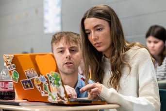 Image of a teacher kneeling beside a student's desk looking at a laptop with a student